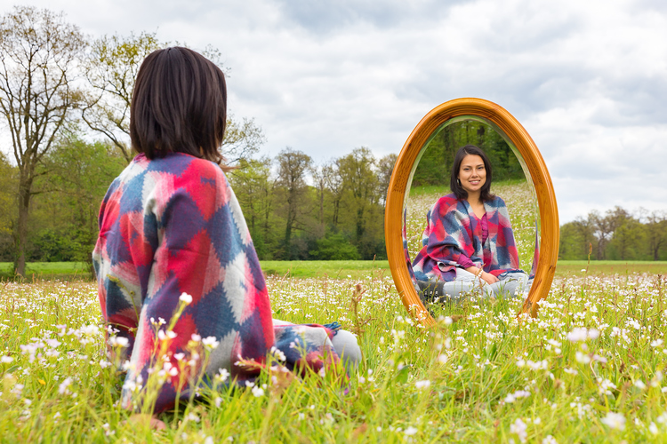 Young colombian woman sitting with mirror in flowering pasture. The woman sits in front of a wooden mirror in a blooming meadow. The pretty person is looking at the reflection of her mirror image. I put the wooden mirror in the middle of a flowering pasture in the netherlands. In spring season you can find several fields of white flowers. The handsome woman is smiling and enjoying nature. In this landscape you can see trees and a horizon in the background.